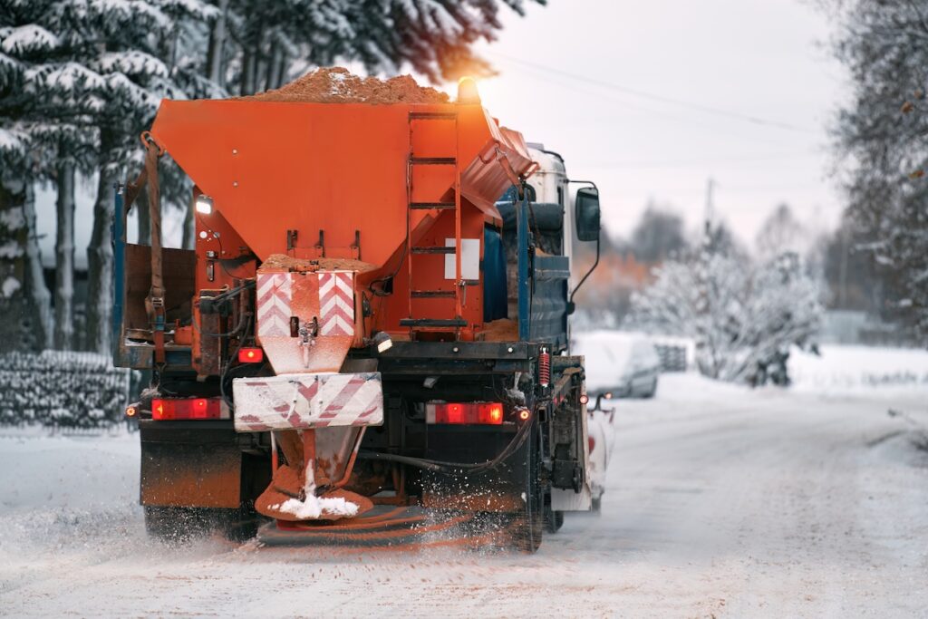 Winter road maintenance snow plow spreading salt and sand mixture on icy pavement to prevent snow buildup.